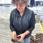 ICE Chef Kathryn Gordon, a woman in a black jacket with short hair and pink glasses, holds up a brown oyster on a dock