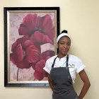 ICE graduate Onika Brown, a woman with black hair pulled back with a white headband wearing a white chef's coat and a grey apron, smiles in front of a framed painting of red flowers