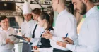 A group of restaurant servers and a chef standing in a line during a training session, taking notes on notepads and clipboards.