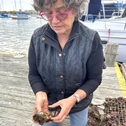 ICE Chef Kathryn Gordon, a woman in a black jacket with short hair and pink glasses, holds up a brown oyster on a dock