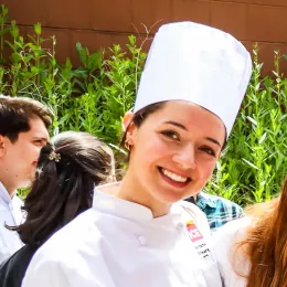 ICE graduate Maddy Devita, a woman with brown hair and brown eyes, smiles in a white chef's coat at ICE's 2023 commencement ceremony