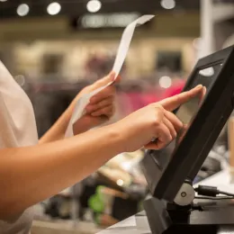 Close-up of a person's hand using a point-of-sale touchscreen monitor while holding a paper receipt.
