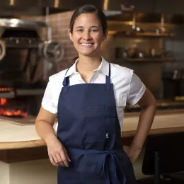 Suzanne Cupps, a brunette woman with her hair in a ponytail, wears a white collared shirt and a blue apron and smiles in front of an open kitchen with a wood-fired stove