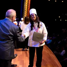 ICE culinary school graduate receives culinary arts diploma wearing chef toque and shaking hand of school president.