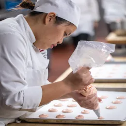 A student pipes pink meringues onto a lined sheet tray