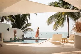 a woman in a hat and pink beach cover up walks along a pool deck with the ocean behind her