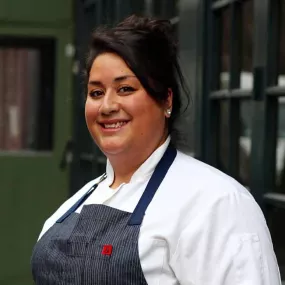 ICE Alumni Ayesha Nurdjaja smiles at the camera in a white chef's coat and black striped apron outside a green building