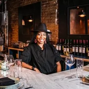 ICE Alumni Shenarri Freeman stands behind the marble counter at Cadance restaurant in a black chef's jacket and black hat