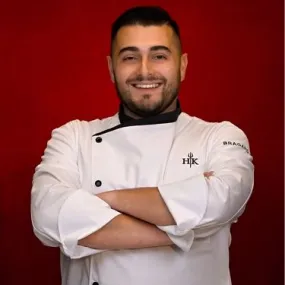 Portrait of ICE alum Chef Shant Halajian smiling warmly with arms folded, wearing a professional white chef coat with a black collar, set against a dark red studio backdrop.