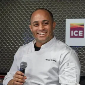 ICE Alumni Michael Jenkins holds a microphone wearing a chef jacket, in front of an ICE logo