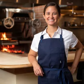 ICE Alumni Suzanne Cupps smiles at the camera while wearing a blue apron and short-sleeved chef jacket. She is leaning on a wooden counter with a flaming wood-burning oven behind her