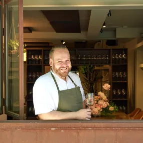 ICE Alumni Samuel Clonts wears an olive green apron and holds a wine glass standing in the open window of his restaurant, 63 Clinton