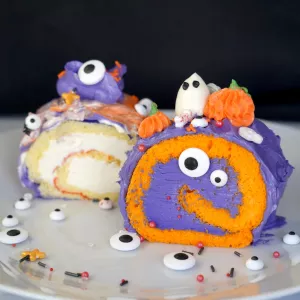 Slices of a purple and orange and white and yellow roulade cake with buttercream pumpkins, ghosts and sprinkles on a white plate