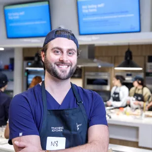 Dr. Nate Wood, a man with dark hair, blue eyes and a backwards blue baseball cap, smiles in a hospital teaching kitchen with his arms crossed