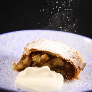 A piece of apple strudel sits next to whipped cream on a white plate
