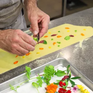 Hands pressing colorful leaves of herbs and petals of flowers on pasta dough