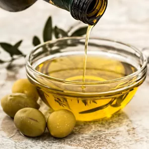 A bottle of olive oil pours into a glass bowl surrounded by green olives