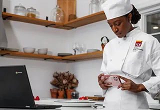 Culinary student in white chef coat and hat stands in home kitchen and photographs dish in front of laptop