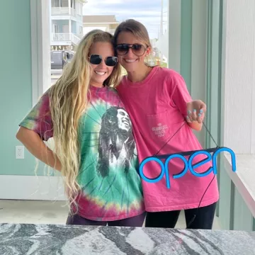 Anna Beth and Lauren stand together smiling and holding an "open" sign