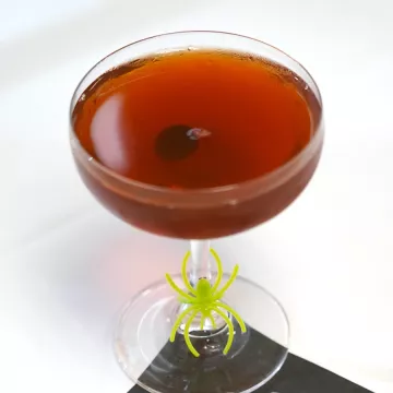 A Black Manhattan cocktail, a dark brown drink with a cherry at the bottom, sits in a coupe glass on a white table with a green plastic spider ring around the stem of the glass