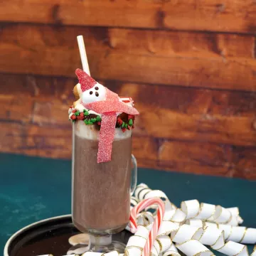 A clear mug of hot chocolate decorated with red and green holiday sprinkles and topped with a marshmallow shaped like a snowman wearing a red gumdrop hat