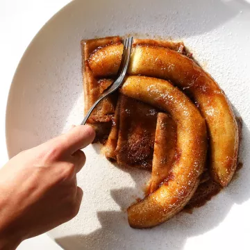 A hand cuts into crepes with caramelized bananas on top, all on a white plate