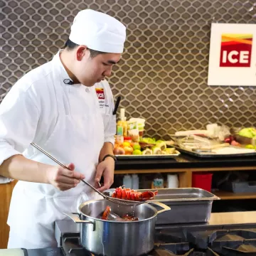 An ICE student in chef's whites holding a netted metal spoon with a lobster tail on it