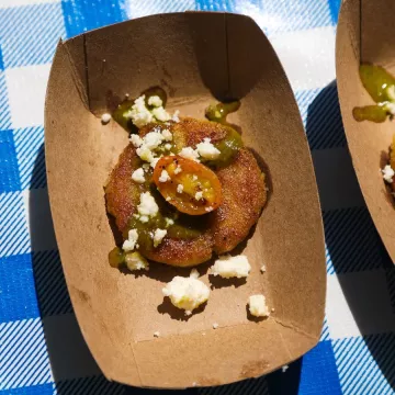 A corn arepa topped with tomato and cheese sits on a cardboard plate on a blue and white checkered background