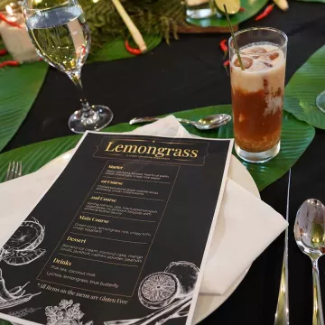 A black restaurant menu with white text sits on a table surrounded by silverware and drinks