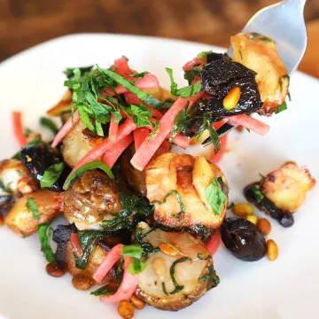 Sunchokes, also known as jerusalem artichokes, are used in a jerusalem artichoke recipe mixed with green herbs, olives and parsnips on a white plate on a wooden table