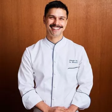 ICE Alumni Orlando Soto smiles at the camera wearing his Le Bernardin chef jacket, against a wood paneled wall