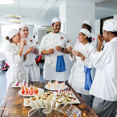 Six ICE students in chef whites gather around a wooden table set with numerous desserts.