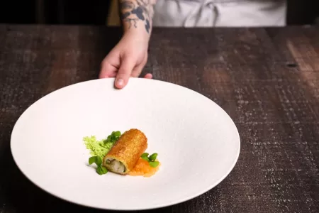 A hands pushes forward a white plate with a golden brown fried roll plated with greens on the side 