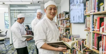 Culinary school student wearing white chef's jacket and hat looks at cookbook in ICE library