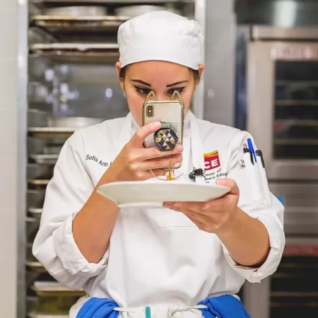 ICE student holds plate with dessert in one hand while taking photo of plate with iphone in other hand.