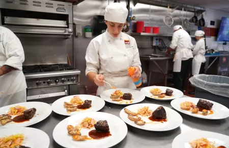 An ICE student in chef's whites plates orange sauce on a plate with brown meat behind many other plated dishes of the same dish