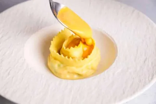 someone out of frame uses a spoon to delicately place an orange puree into the folds of a circular pasta