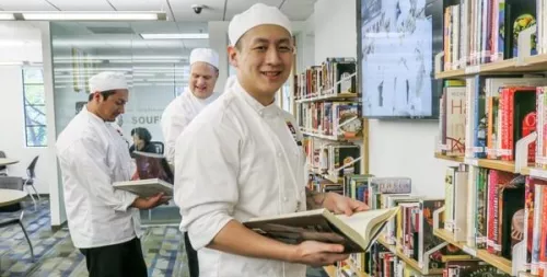 Culinary school student wearing white chef's jacket and hat looks at cookbook in ICE library