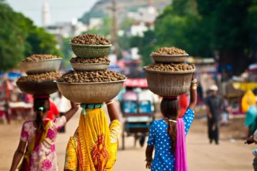 three women in colorful saris carry buckets of nuts on their heads down a busy street