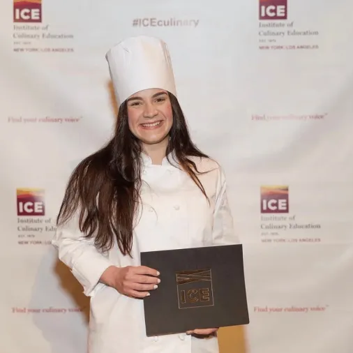 Maried Arroyo smiling in a white chef uniform and hat, holding her Institute of Culinary Education diploma in front of an ICE step-and-repeat backdrop.