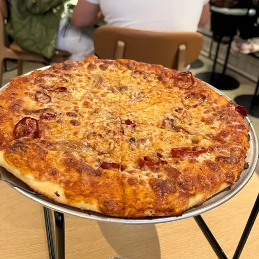 A cheesy pizza on a metal tray sits on a metal pizza holder stand on a wooden table