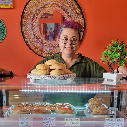 Chef Nilüfer Goodson, a woman wearing a green shirt and glasses, smiles behind a pastry case at her eponymous restaurant