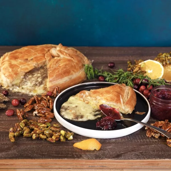 A slice of baked Brie cheese wrapped in baked puff pastry sits on a black plate in front of a wheel of the baked Brie cheese. They are both surrounded by green herbs, orange halves, candied nuts and cranberries