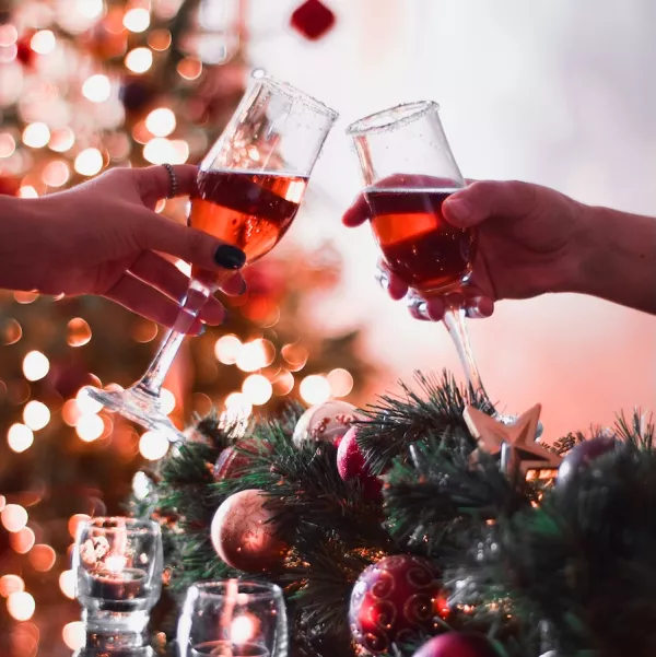 Two hands clink wine glasses with red liquid together above a table with garland and ornaments on it