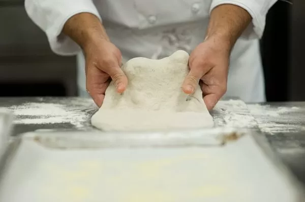 Close-up shot of chef's hands stretching bread dough on a stainless steel countertop. 