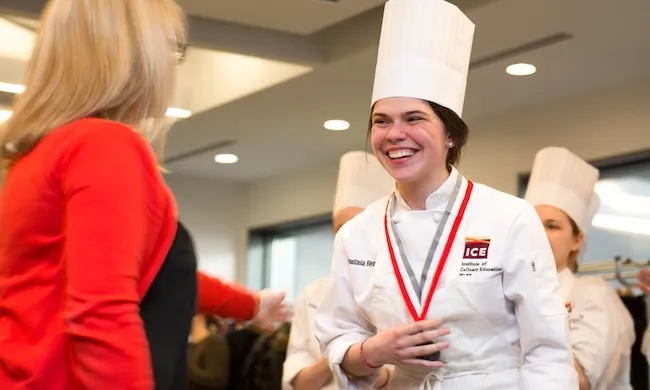 An Institute of Culinary Education graduate in chef whites smiles as she shakes the had of an instructor
