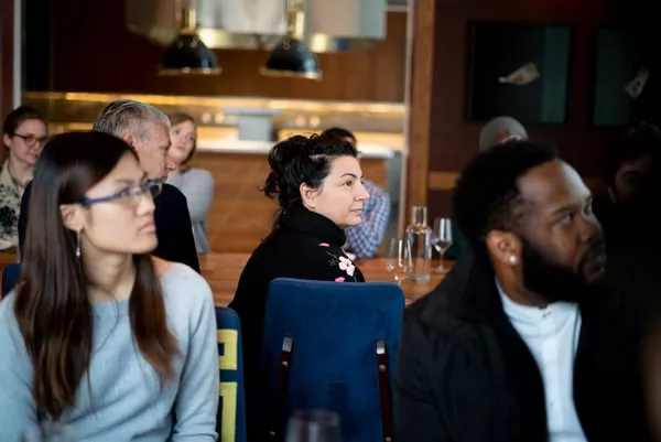 Tourism, Travel & Hospitality students of various ages and races sit at tables in a restaurant and look to the right at a speaker outside of the frame