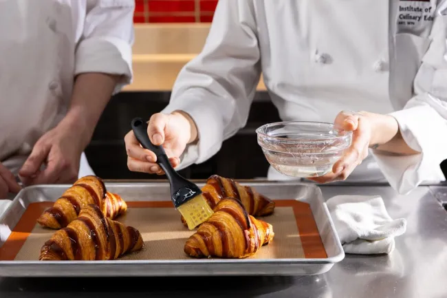 an Advanced Pastry & Baking Technique student brushes a glaze onto fresh croissants