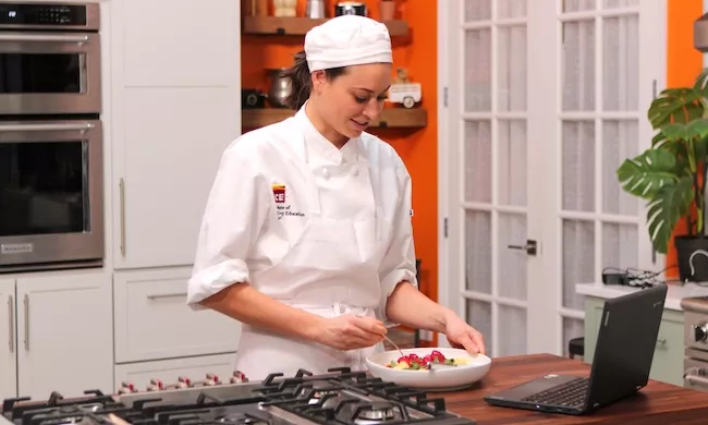 An ICE online culinary associate degree student in chef's whites plates a dish while looking at a laptop