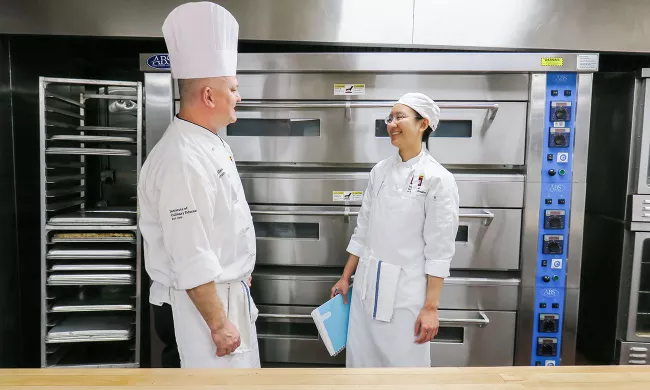 A pastry student and a chef instructor stand in front of industrial deck ovens at ICE